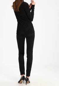 Donna con lunghi capelli castani, che indossa un top nero a maniche lunghe, pantaloni skinny neri e scarpe con tacco alto nere, in piedi e girata di spalle.