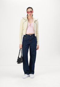 Giacca crema con zip sopra un crop top a coste rosa chiaro. Jeans a gamba larga blu scuro. Sneakers bianche e una borsa nera con dettagli argentati. Occhiali da sole rosa.