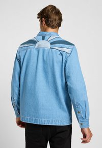 Ljusblå denimjacka med en soldesign på baksidan och kontrasterande blå ränder. Inkluderar knappmanschetter och en klassisk krage.