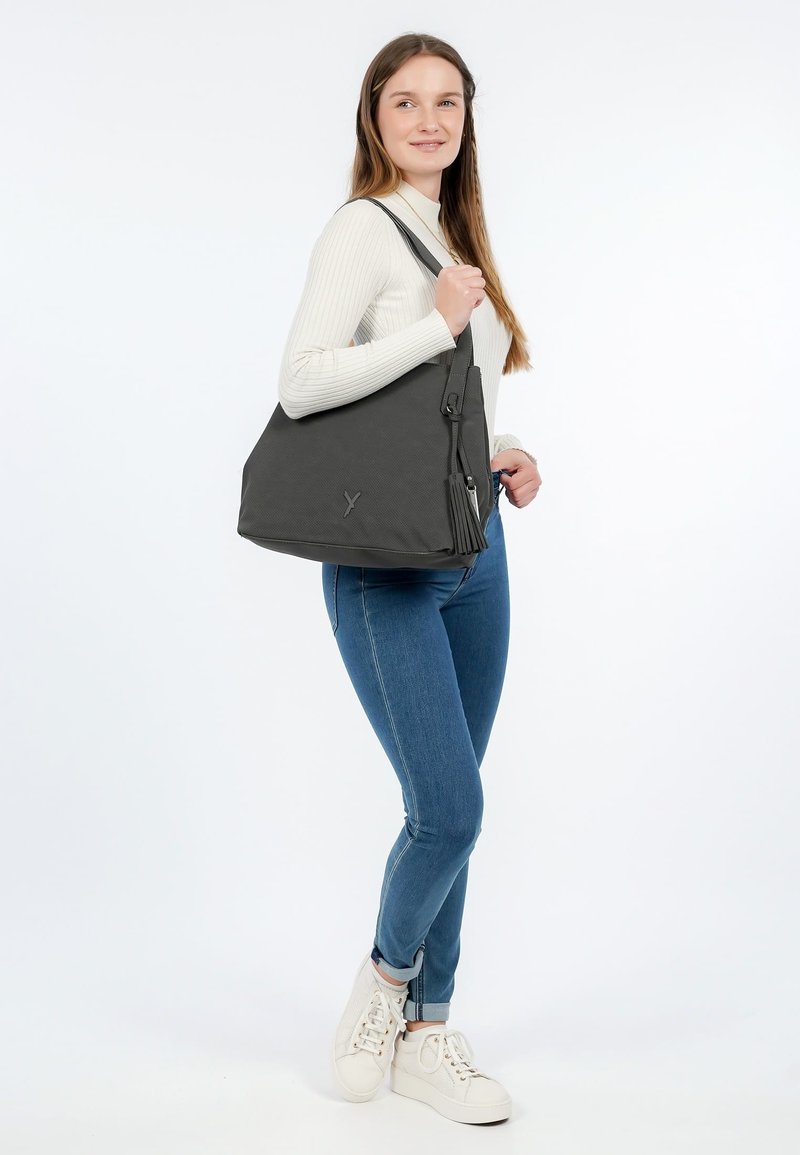 Donna che indossa un maglione bianco e jeans blu, tiene una grande borsa a tracolla grigio scuro con dettagli a nappine, in piedi su sfondo bianco.