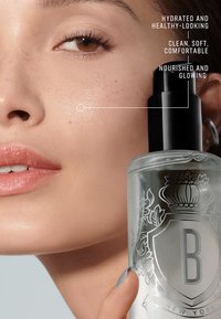 Heldere glazen bottle met zwarte pompdeksel, voorzien van verfijnde gravures. De huid van het model ziet er glad uit, met een natuurlijke glans en subtiele sproeten.
