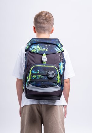 Child seen from behind wearing a black and blue splattered backpack with a soccer ball badge and cleat keychain.