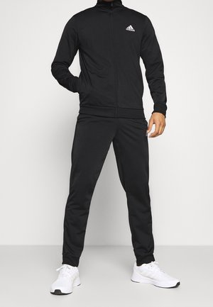 Homme portant un survêtement Adidas noir avec des baskets blanches, debout devant un fond clair uni, les mains relâchées le long du corps.