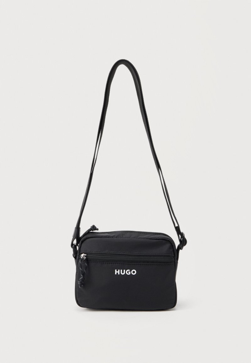 Μαύρη ορθογώνια τσάντα ώμου με εμπρόσθια τσέπη με φερμουάρ, λογότυπο "HUGO" και ρυθμιζόμενο λουρί σε λευκό φόντο.