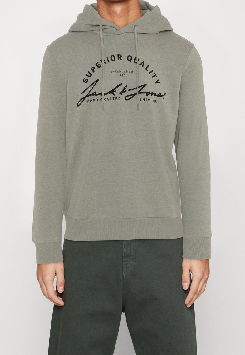Sweat à capuche vert avec cordon de serrage, arborant un logo imprimé noir et un texte sur le devant. Le matériau semble doux et texturé, de style décontracté.