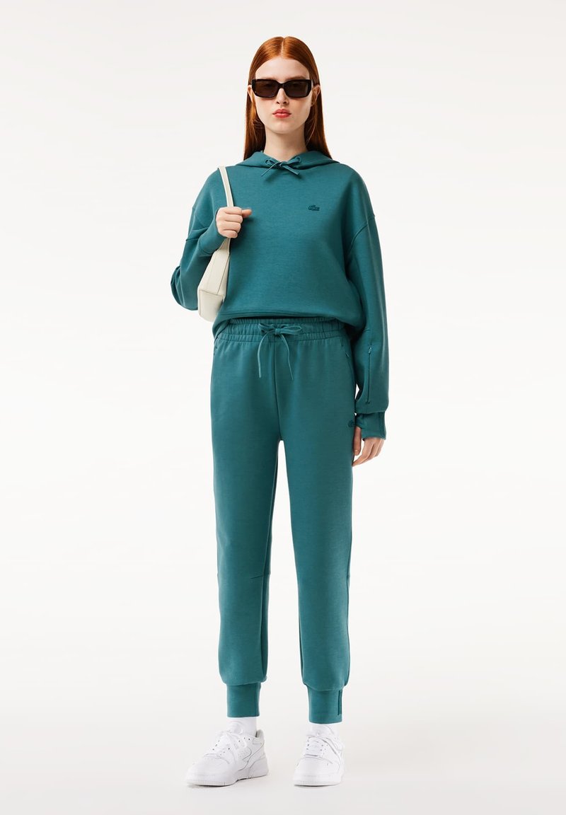Turkusowa bluza z kapturem i sznurkiem, zestawiona z dopasowanymi, zwężającymi się dresami. Model nosi białe sneakersy i trzyma małą beżową torbę.
