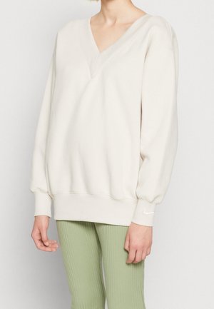 Person trägt ein locker sitzendes, cremeweißes V-Ausschnitt-Sweatshirt mit langen Ärmeln und hellgrünen, gerippten Hosen vor einem einfarbigen Hintergrund.