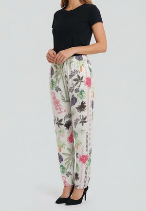Femme portant un haut noir à manches courtes, un pantalon blanc avec des motifs floraux et feuillus colorés, et des chaussures noires à talons hauts, debout de profil.