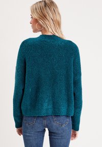 Suéter de color azul verdoso, suave al tacto, con un corte relajado, escote redondo y mangas largas, combinado con jeans azules. Textura suave y dobladillo cortado.
