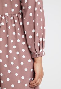 Šaty s dlouhým rukávem z látky v barvě starorůžové s bílými puntíky, se stahovaným pasem a zapínáním na knoflíky na manžetách. Hladká textura.