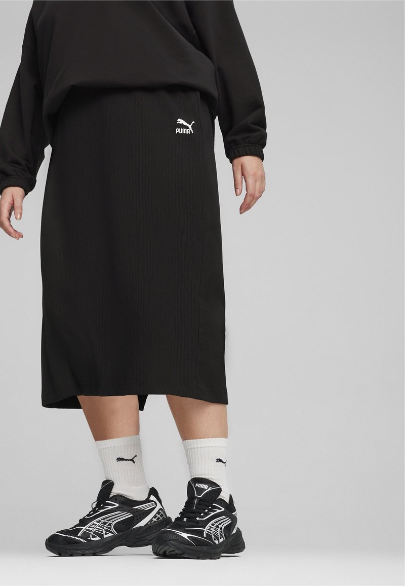 Svart ribbat stickat kjol med elastisk midjeband och en vit Puma-logotyp. Kombination med svarta och vita löparskor och vita strumpor.