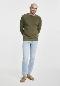 Oliwkowy sweter dzianinowy z teksturowanej tkaniny, jasnoniebieskie dżinsy i tawne mokasyny. Model stoi z rękami złożonymi za plecami.