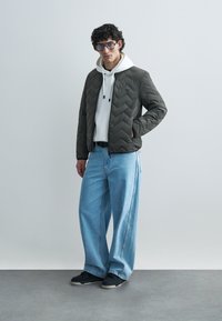 Padded olive green jacket with a chevron pattern, worn over a white hoodie; paired with wide-legged light blue jeans and black sneakers.