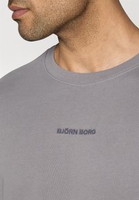 Šedé tričko s okrúhlym výstrihom vyrobené z hladkej bavlny, s tmavomodrým vyšívaným logom "BJÖRN BORG" na hrudi. Detailný záber na textúru tkaniny.