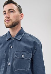 Mørkeblå skjorte med knapper fremme, lavet af bomuld med en standard krave, to brystlommer og kontrasterende hvide syninger.