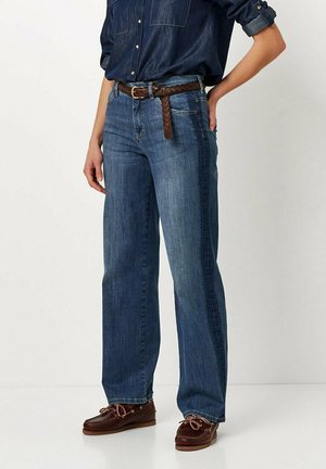 Denim-Jeans in mittelblau mit lockerer Passform, ausgestattet mit einem geflochtenen braunen Ledergürtel, und kombiniert mit braunen Bootsschuhen.