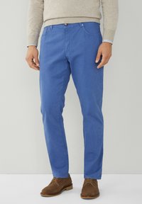 Muškarac u svijetloplavim slim-fit hlačama, bež puloveru i smeškim zamširanim cipelama sa vezicama, stoji protiv jednostavne svijetle pozadine.