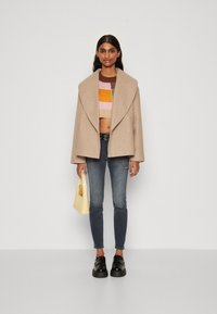 Μπεζ παλτό από μίγμα μαλλιού με φαρδιές πέτες, πολύχρωμο κοντό πλεκτό πουλόβερ, μπλε skinny τζιν, μαύρα παπούτσια και μία μικρή κίτρινη τσάντα.