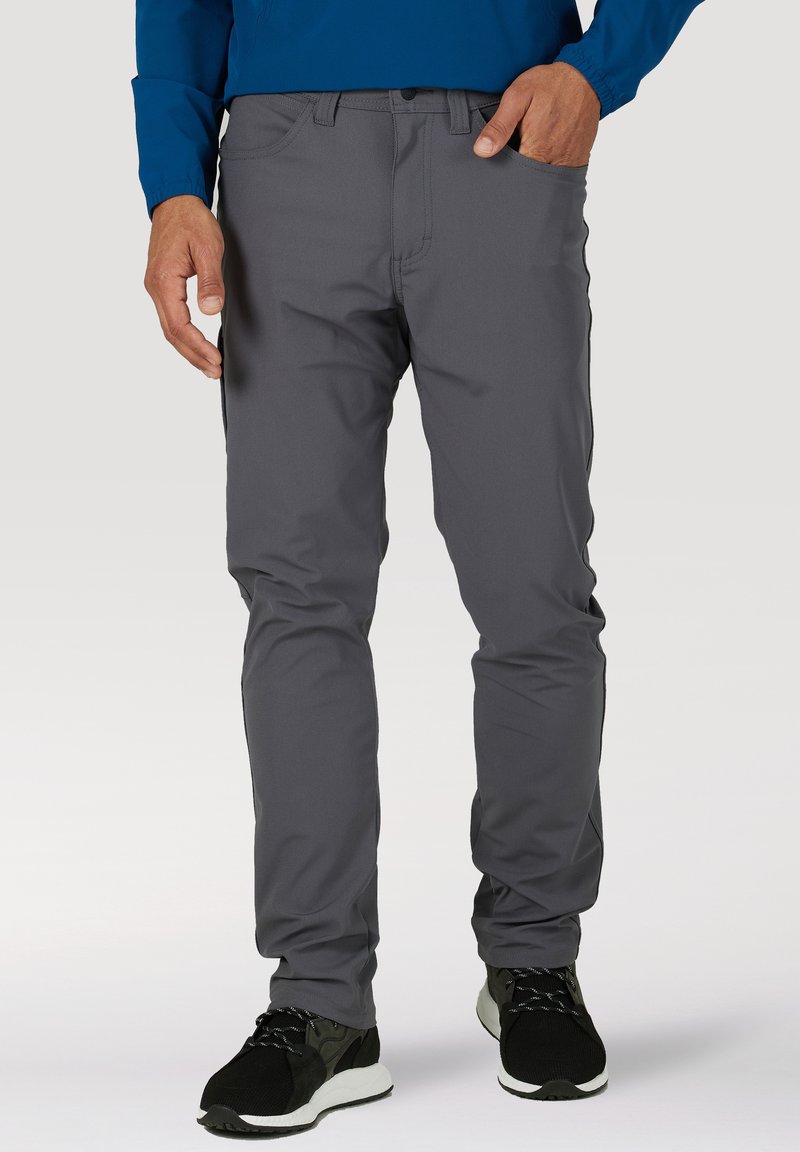 Mann trägt graue Hose und schwarze Turnschuhe, steht mit einer Hand in der Tasche vor schlichtem Hintergrund.