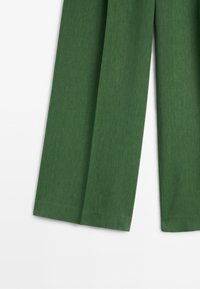 Twee brede groene broekspijpen van gestructureerde stof tegen een witte achtergrond.