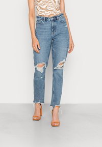 Vaqueros desgastados de un azul claro con dobladillos deshilachados y roturas en las rodillas, presentan un diseño clásico de cinco bolsillos. Llevados con sandalias de tacón naranjas.