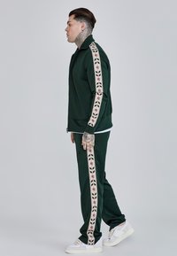 Zelený tréningový súprava s bundou na zips a bočnými prúžkami s kvetinovým vzorom. Štruktúrovaná tkanina doplnená bielymi teniskami.