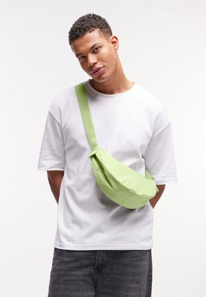 Giovane uomo con capelli corti e ricci che indossa una maglietta bianca e una borsa a tracolla verde chiaro, in piedi davanti a uno sfondo bianco uniforme.