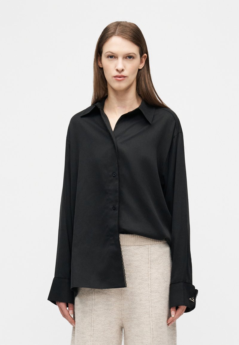 Γυναίκα με ίσια καστανά μαλλιά, φορώντας μια υπερμεγέθη μαύρη πουκάμισα με κουμπιά και ανοιχτό μπεζ πλεκτό παντελόνι σε απλό λευκό φόντο.