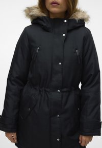 Melnā parka ar mākslīgo kažokādu iestrādātu kapuci, ar rāvējslēdzēju krūšu kabatām, pogām ar pogu un regulējamu jostasvietu. Gluda auduma ar matētu apdari.