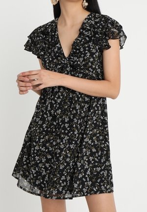 Robe noire à fleurs avec décolleté en V volanté et manches papillon portée par une femme aux longs cheveux et boucles d'oreilles en or.