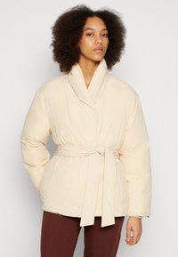Casaco puffer bege com um design envolvente, gola xale e cintura com cinto. O tecido apresenta uma textura suave e bolsos laterais.