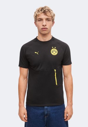 Jeune homme aux cheveux blonds courts portant un t-shirt noir Puma avec un logo BVB 09 jaune et une poche zippée jaune, associé à un jean bleu.
