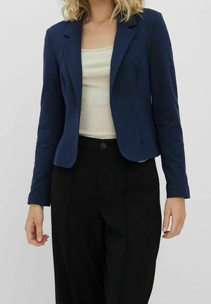 Donna che indossa un blazer blu navy, top crema senza maniche e pantaloni neri a vita alta su uno sfondo chiaro uniforme.