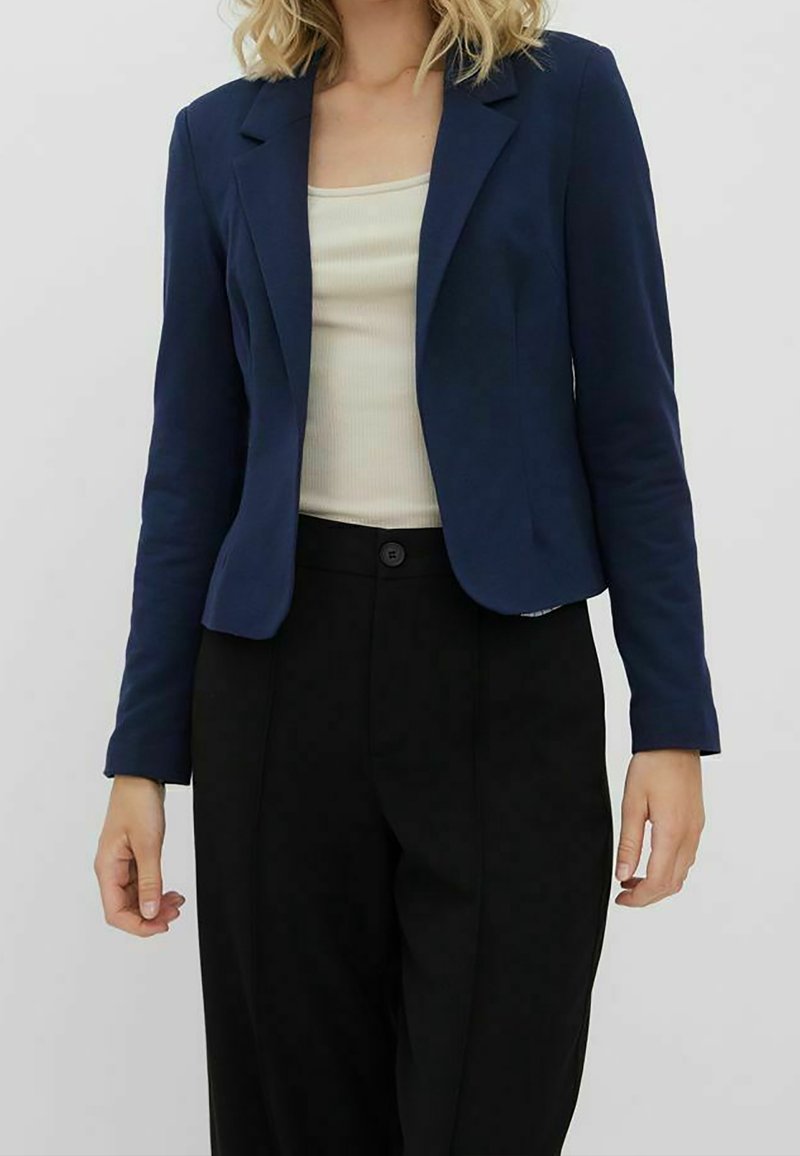 Femme portant un blazer bleu marine, un top crème sans manches et un pantalon noir taille haute sur un fond clair uni.