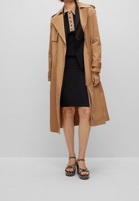 Vaaleanruskea trench-coat mustan mekon päällä, jossa on erottuva kaulus. Mustat plattopohjaiset kengät, joissa on kuvioitu design, täydentävät asua.