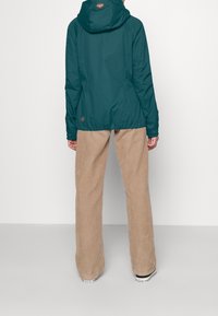 Tyrkysová voděodolná bunda s kapucí, elastickými manžetami a šňůrkou na spodním lemu; v kombinaci s béžovými kordovými kalhotami a černými teniskami.