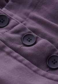 Lila tyg med tre svarta fyrhålsknappar sydda i en vertikal rad, som visar detaljerad sömnad och textur.