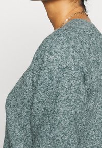 Tummanvihreä neulottu villapaita, jossa on teksturoitu kuvio, pyöreä pääntie ja väljä istuvuus, sekä pudotetut olkapäät ja pehmeä kangas.