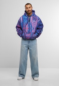 Uomo in piedi con le mani in tasca, che indossa un hoodie a motivi blu e viola, jeans leggeri a gamba larga di colore azzurro e sneakers bianche su sfondo neutro.