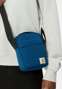 Bolso cruzado de tela azul con correa ajustable, bolsillo frontal con cremallera y parche de logotipo; forma cuadrada con textura suave y diseño resistente.