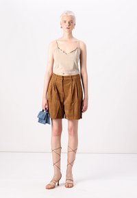 Un top beige court avec un décolleté festonné, un short plissé marron et des talons beiges à lanières. Tenant un petit sac à main bleu.