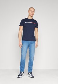 Marineblauw t-shirt met korte mouwen met een horizontale witte, rode en blauwe streep over de borst, gecombineerd met lichtblauwe jeans en sneakers.