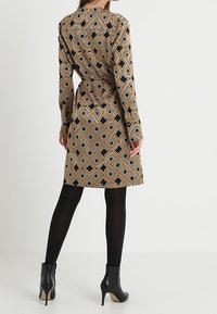 Langarmkleid mit geometrischem Muster in Braun und Schwarz, das über einen Gürtel und einen geschwungenen Saum verfügt. Kombiniert mit schwarzen Strumpfhosen und high-heeled Stiefeln.