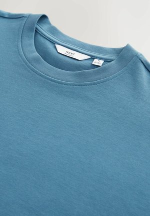 T-shirt en coton bleu clair avec un col ras du cou, texture lisse et étiquette indiquant "NEXT, coupe décontractée" visible à l'intérieur.