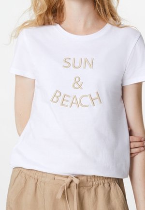 Biała koszulka z beżowym haftowanym napisem "SŁOŃCE I PLAŻA" noszona z beżowymi spodniami na sznurku przez osobę o jasnej karnacji i falujących blond włosach.