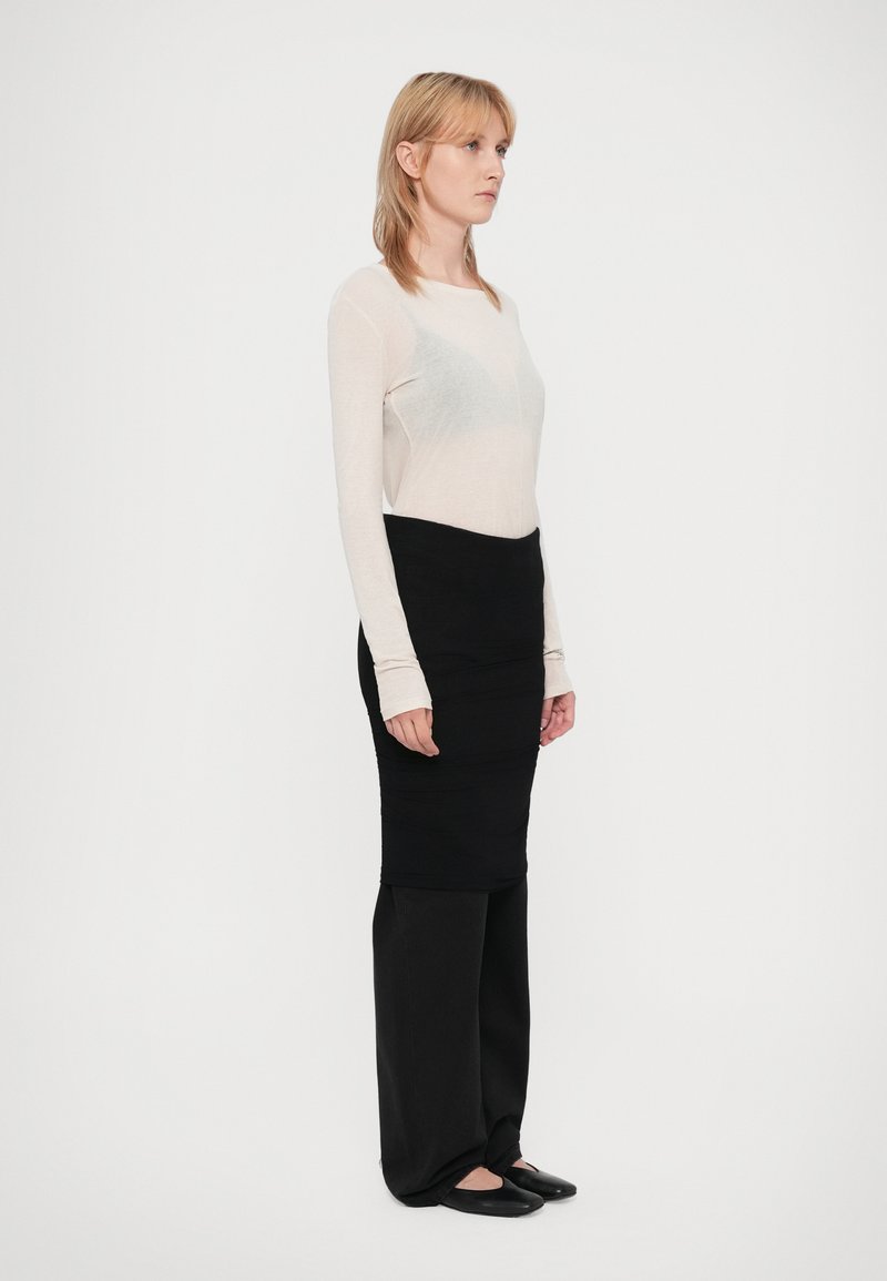 Eine Frau mit blonden Haaren steht seitlich, trägt ein langärmliges, durchscheinendes weißes Oberteil, einen schwarzen Rock über schwarzen Hosen und schwarze Ballerinas.