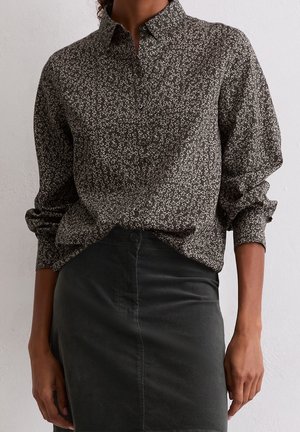 Donkerbruin overhemd met knopen, voorzien van een klein wit bloemmotief, ingestopt in een hoog getailleerde zwarte rok, met de mouwen opgerold tot de onderarmen.