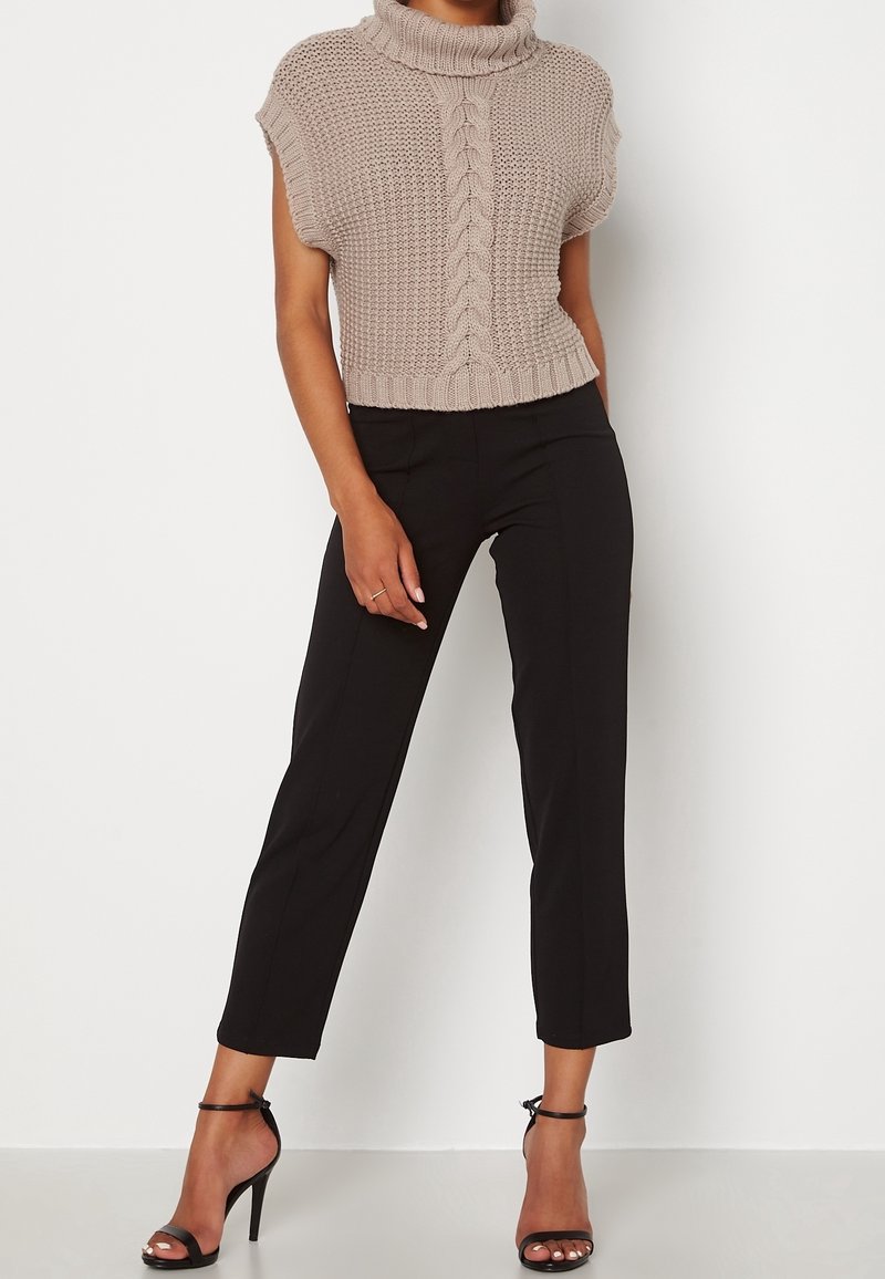 Top court beige en maille avec un col montant et un motif en torsades, associé à un pantalon noir tailleur et des talons noirs à lanières.