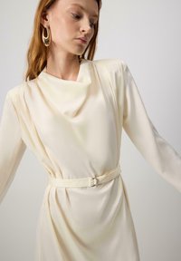 Punane juustega naine kannab suuri kuldseid kõrvarõngaid ning kreemivärvi pika varrukaga kleiti, millel on lokkis kaelus ja vööpais.