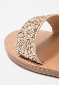 Kulta- ja hopeakiiltoiset liukusandaalit, joissa on beige nahkapohja, kaareva hihna ja sileä pinta. Reunat on siististi ommeltu.